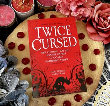 Copy of Twice Cursed, edited by Marie O'Regan and Paul Kane, on a gold cloth decorated with red beads, pink roses and candles, and silver leaves